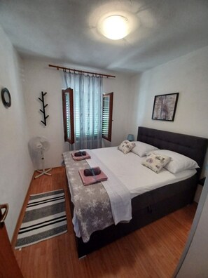 Un dormitorio con cama, ventilador de pie, ventana con cortinas, un cuadro en la pared y suelo de madera.
