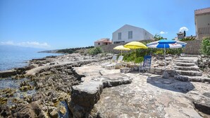 Eine Küstenlandschaft mit einem steinernen Weg, der zum Strand führt, einem weißen Gebäude mit rotem Dach und zwei Sonnenschirmen, die Schatten spenden.