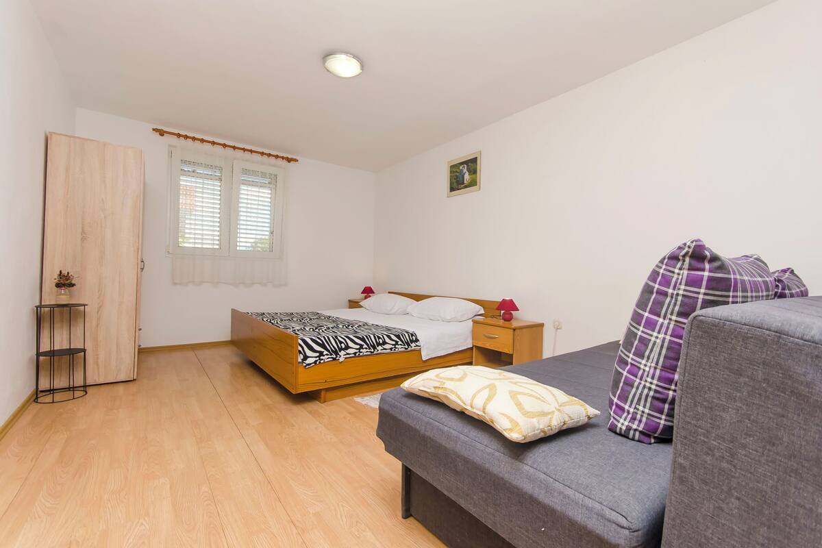 A bedroom with a wooden bed, a grey sofa, a bedside table with a lamp, and a window with blinds.