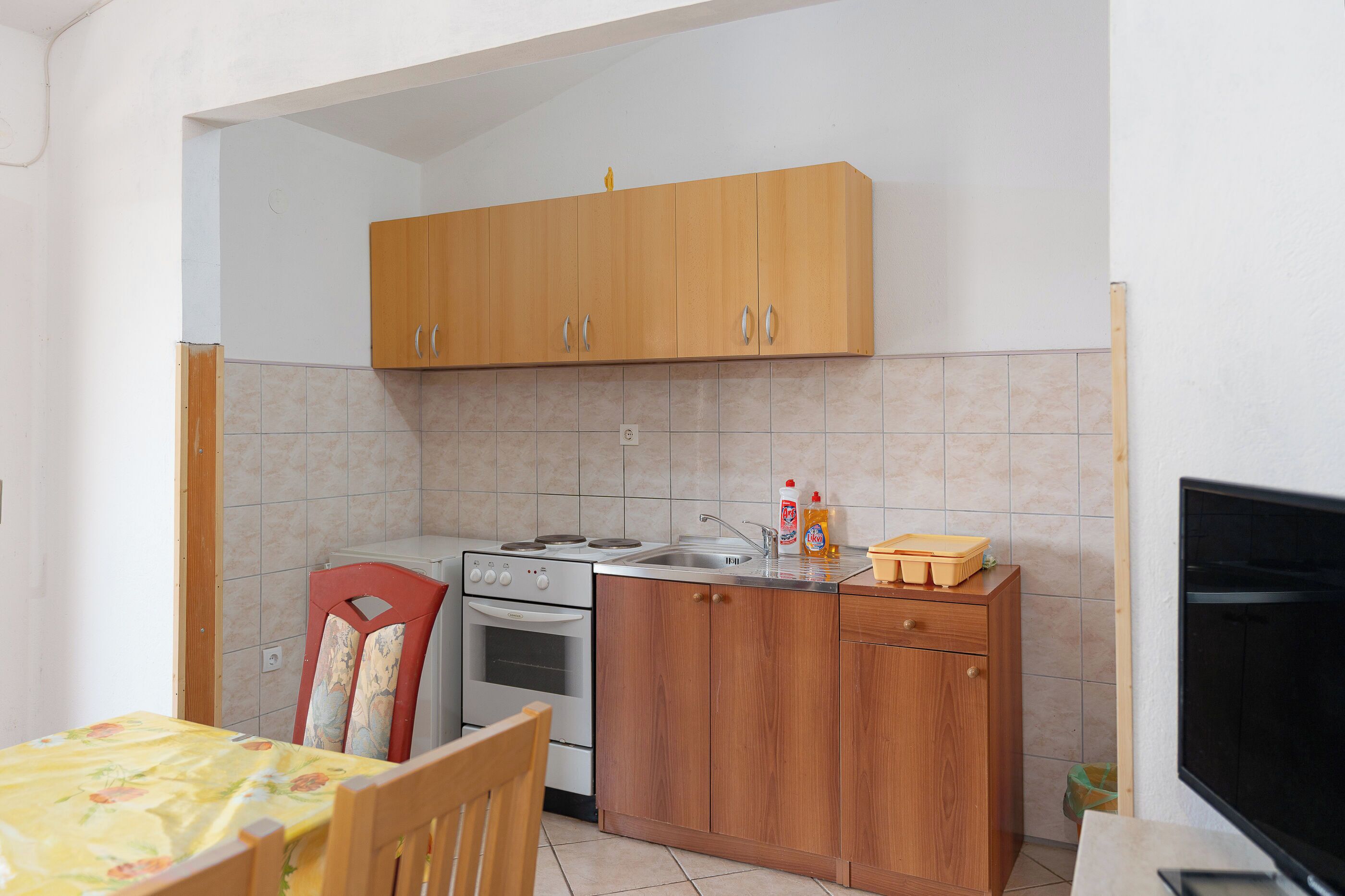 A small kitchen with wooden cabinets, a stove, and a sink. There is a chair and a table with a floral tablecloth.