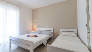 Ein kleines, sauberes Zimmer mit zwei Einzelbetten, einem Nachttisch mit Lampe und einem Fenster mit durchsichtigen Vorhängen.