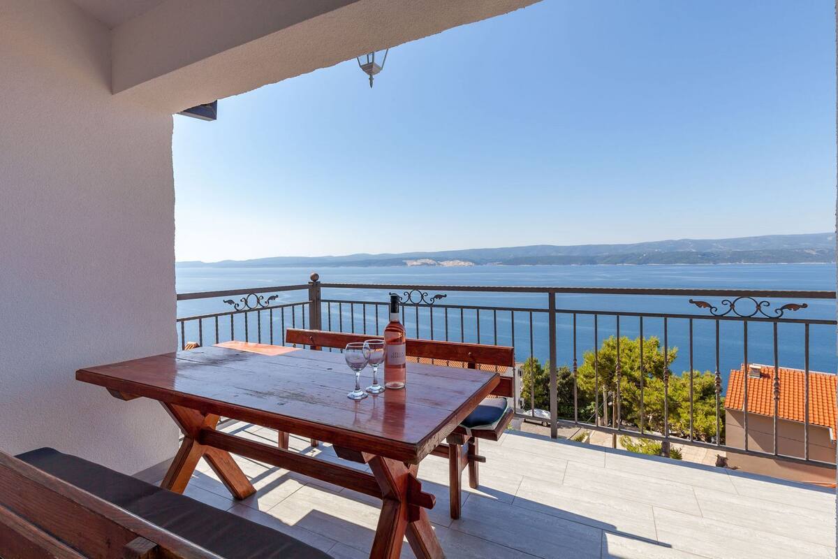 A wooden table with two wine glasses and a bottle on a balcony overlooking a body of water.