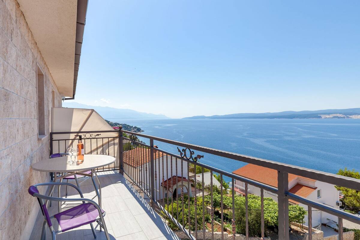 A balcony with a table and chairs, overlooking a body of water and distant mountains.