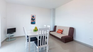 Ein modernes Wohnzimmer mit einem weißen Esstisch, weißen Stühlen, einem braunen Sofa mit einem rot-weißen Kissen, einem Flachbildfernseher auf einem Glastisch und einem Wandbild.