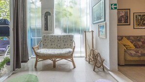 Une pièce avec un fauteuil en osier, un canapé, un tapis vert et une fenêtre avec des rideaux.