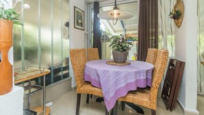 Un coin repas avec des chaises en osier, une table recouverte d’une nappe violette, un luminaire suspendu et une plante en pot.