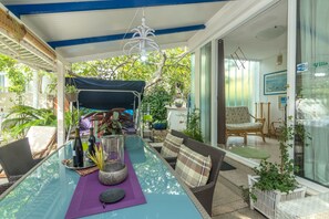 Een overdekt terras met een tafel, stoelen en plantenpotten.