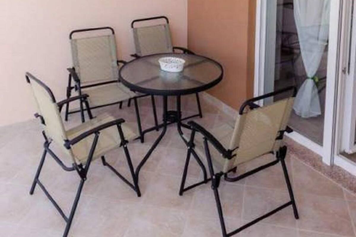 A small outdoor patio with a round table and four chairs, an air conditioning unit, and a sliding glass door with a curtain.