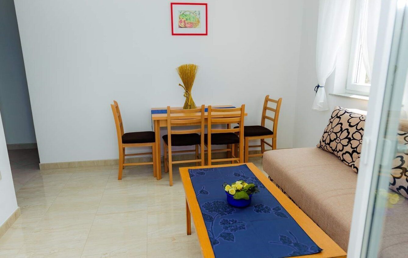 Une petite pièce bien éclairée, avec un lit, une table à manger entourée de chaises et un chemin de table bleu agrémenté d’un arrangement floral.