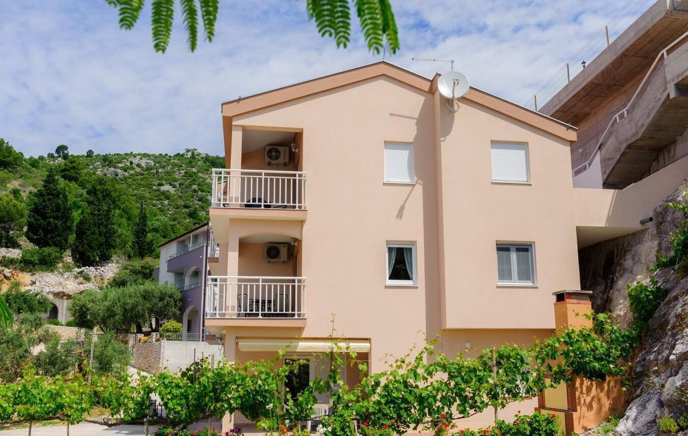 A two-story building with a balcony on the upper floor, surrounded by greenery and a hillside.