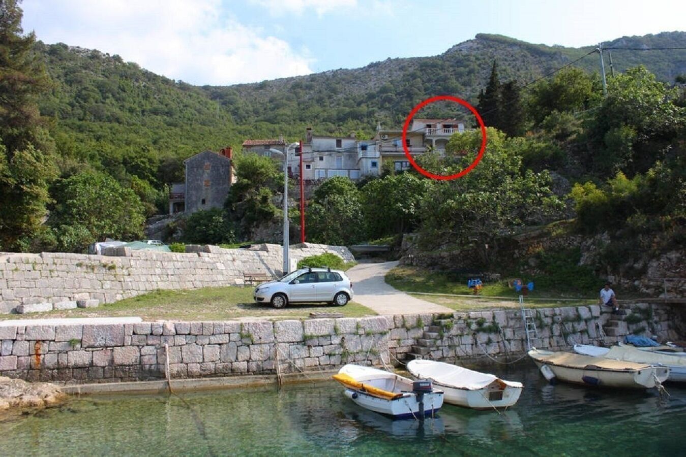 A stone wall, a paved path, and boats in a body of water with a mountainous backdrop.