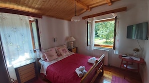 Ein Schlafzimmer mit Holzbalkendecke, einem Bett mit roter Bettdecke, einem Nachttisch mit Lampe, einem Fenster mit Blick auf Grünflächen und einem kleinen Tisch mit einer Vase.