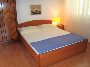 A single bed with a wooden frame and a blue bedspread, a bedside table with a lamp, and a wooden wardrobe. - Apartment Dane (Makarska)