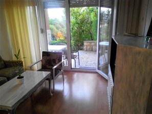 A room with a wooden floor, a sofa, a table with a plant, and a sliding glass door leading to an outdoor area with a table and chairs. - Apartment Dane (Makarska)