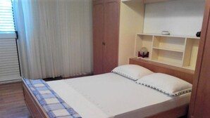 A single bed with a wooden frame, a white mattress, and a quilt. There is a wooden wardrobe to the left and a shelf unit with a mirror above the bed. - Apartment Dane (Makarska)