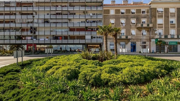 A multi-story building with balconies, a landscaped garden with palm trees, and a visible storefront with a sign. - Apartment Deni (Split)