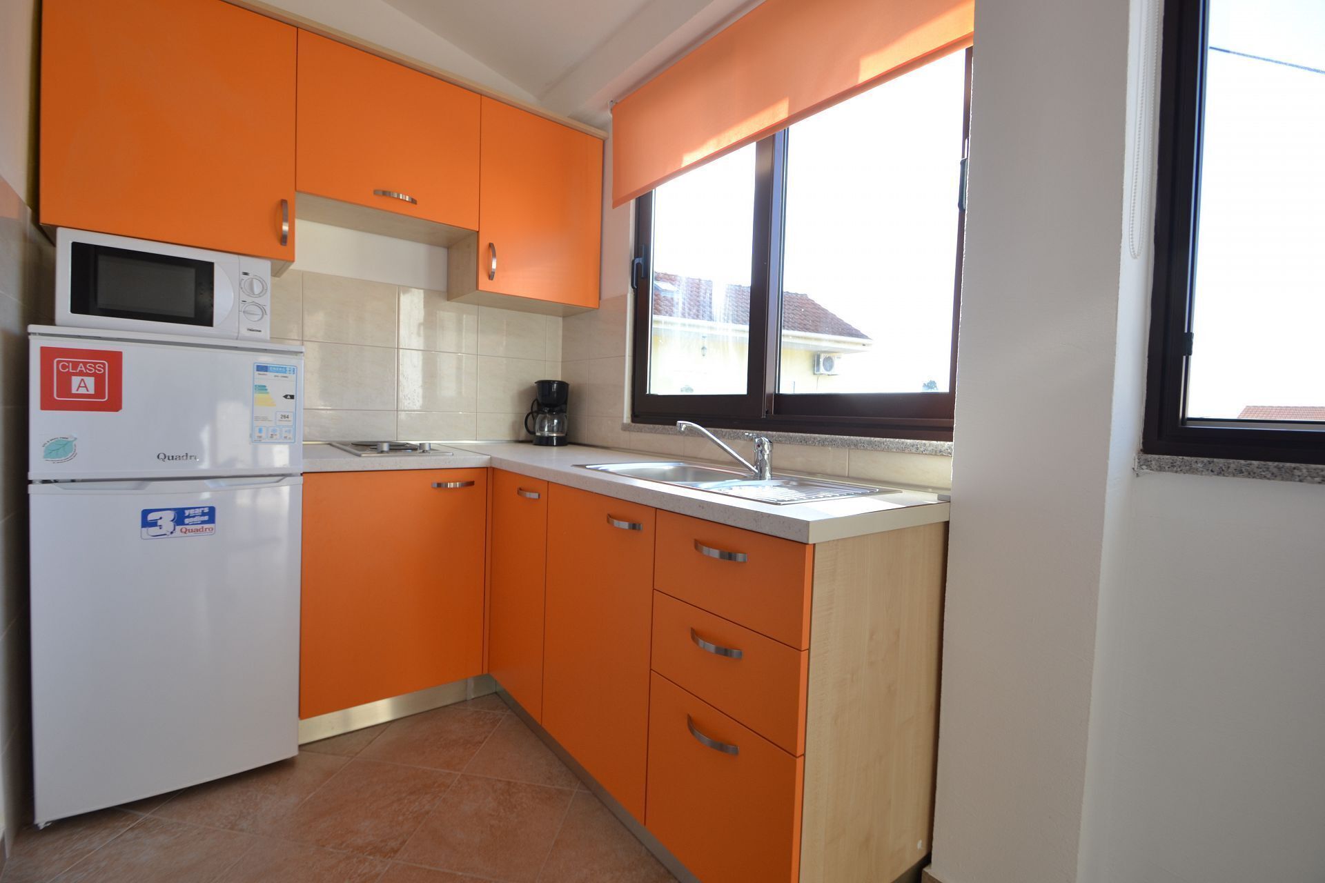 A kitchen with orange cabinets, a white refrigerator, a microwave, and a window with a view of buildings outside.