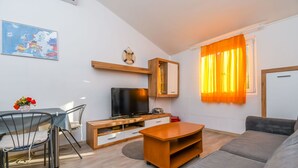 Egy modern nappali, melyben síkképernyős televízió, fából készült bútorok, egy kanapé és egy székekkel körülvett étkezőasztal található.