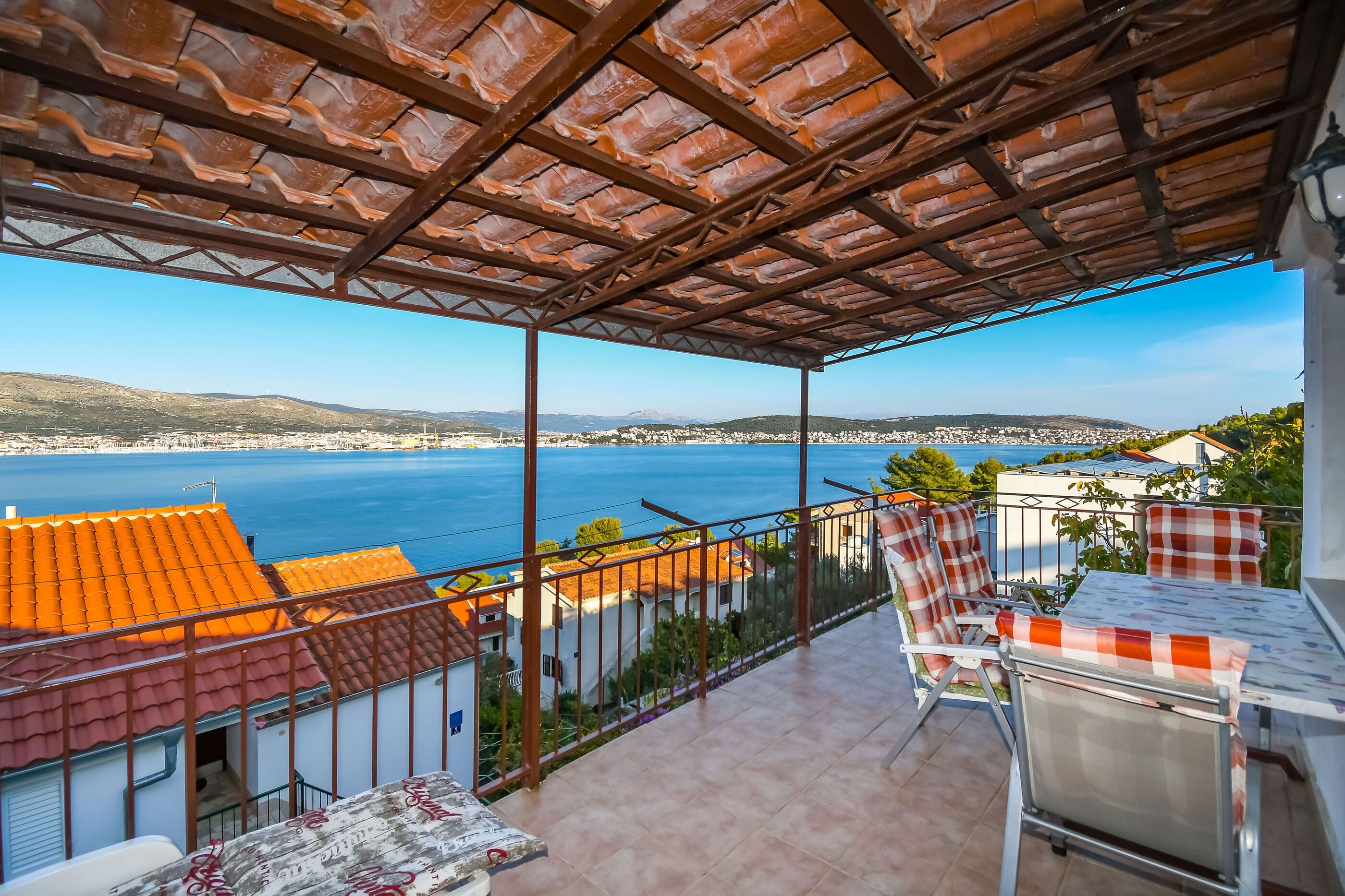 A balcony with a view of the sea, a table with chairs, and a covered area with a tiled roof.