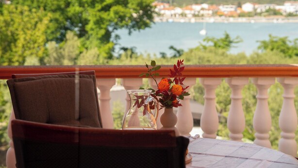 Ein Balkon mit einem Stuhl, einer Blumenvase und Blick auf ein Gewässer sowie Gebäude in der Ferne.