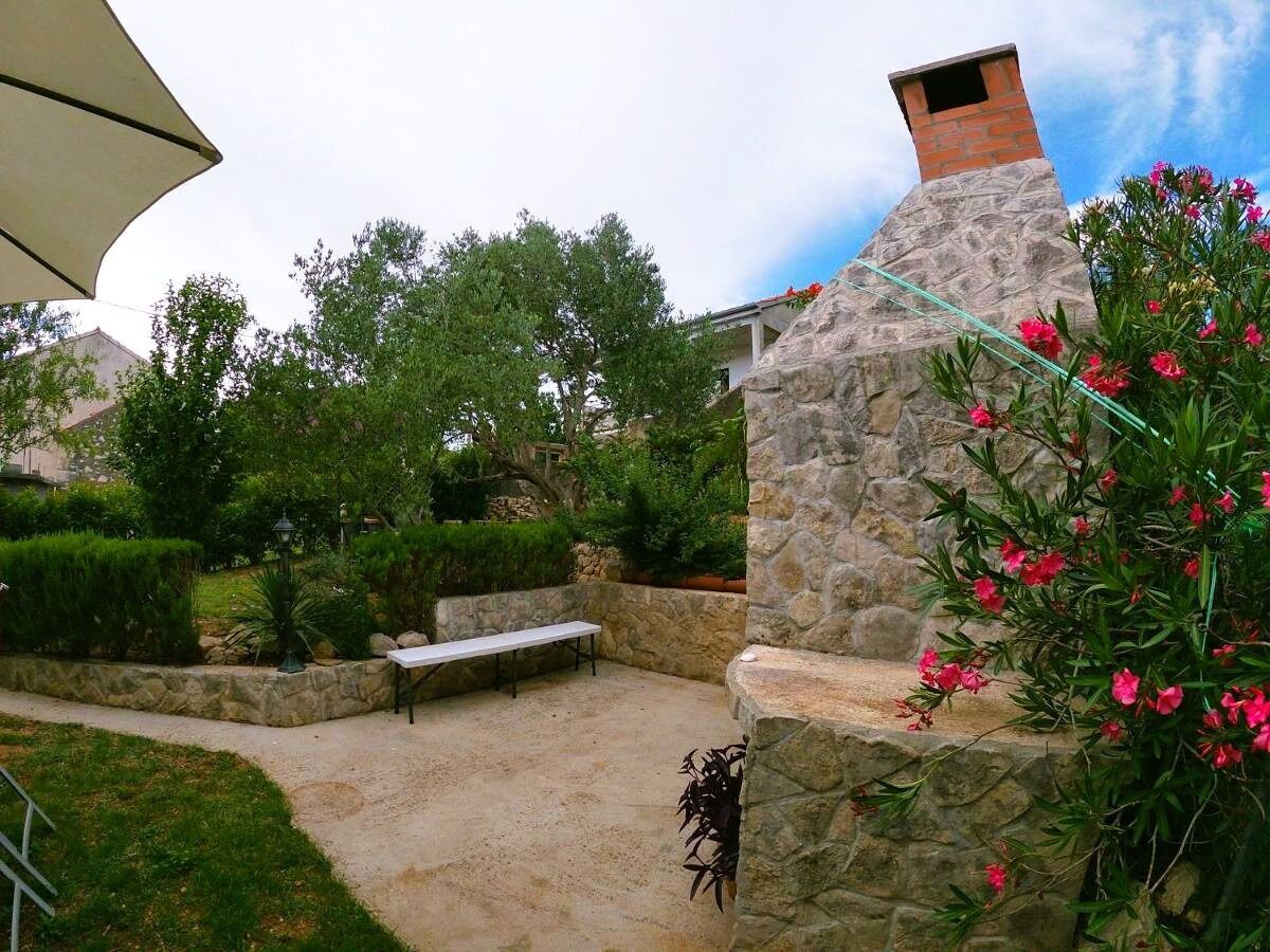 A stone-built outdoor area with a bench and a table, surrounded by greenery and a stone wall.