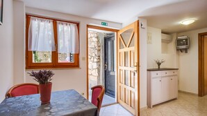 A dining area with a table and chairs, a potted plant, a stone wall, a black door, and a kitchen area with cabinets and a television. - Apartments Stipe (Mali Losinj)