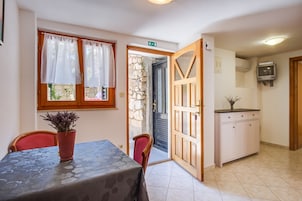 Un comedor con una mesa y sillas, una planta en maceta, un muro de piedra, una puerta negra y una zona de cocina con armarios y un televisor.
