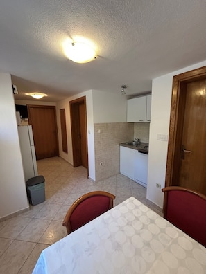 A small kitchen with a refrigerator, sink, and cabinets, a dining table with chairs, and a trash bin. - Apartments Stipe (Mali Losinj)