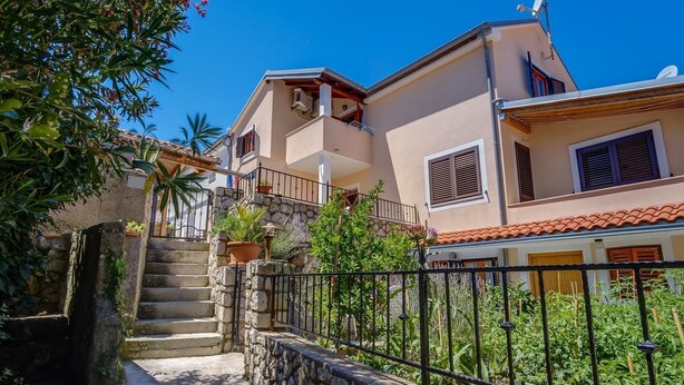 A two-story building with a stone staircase leading to a balcony, surrounded by greenery and a metal fence. - Apartments Stipe (Mali Losinj)