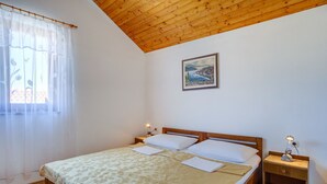 A bedroom with a wooden ceiling, a bed with a floral bedspread, two bedside tables with lamps, and a window with sheer curtains. - Apartments Stipe (Mali Losinj)