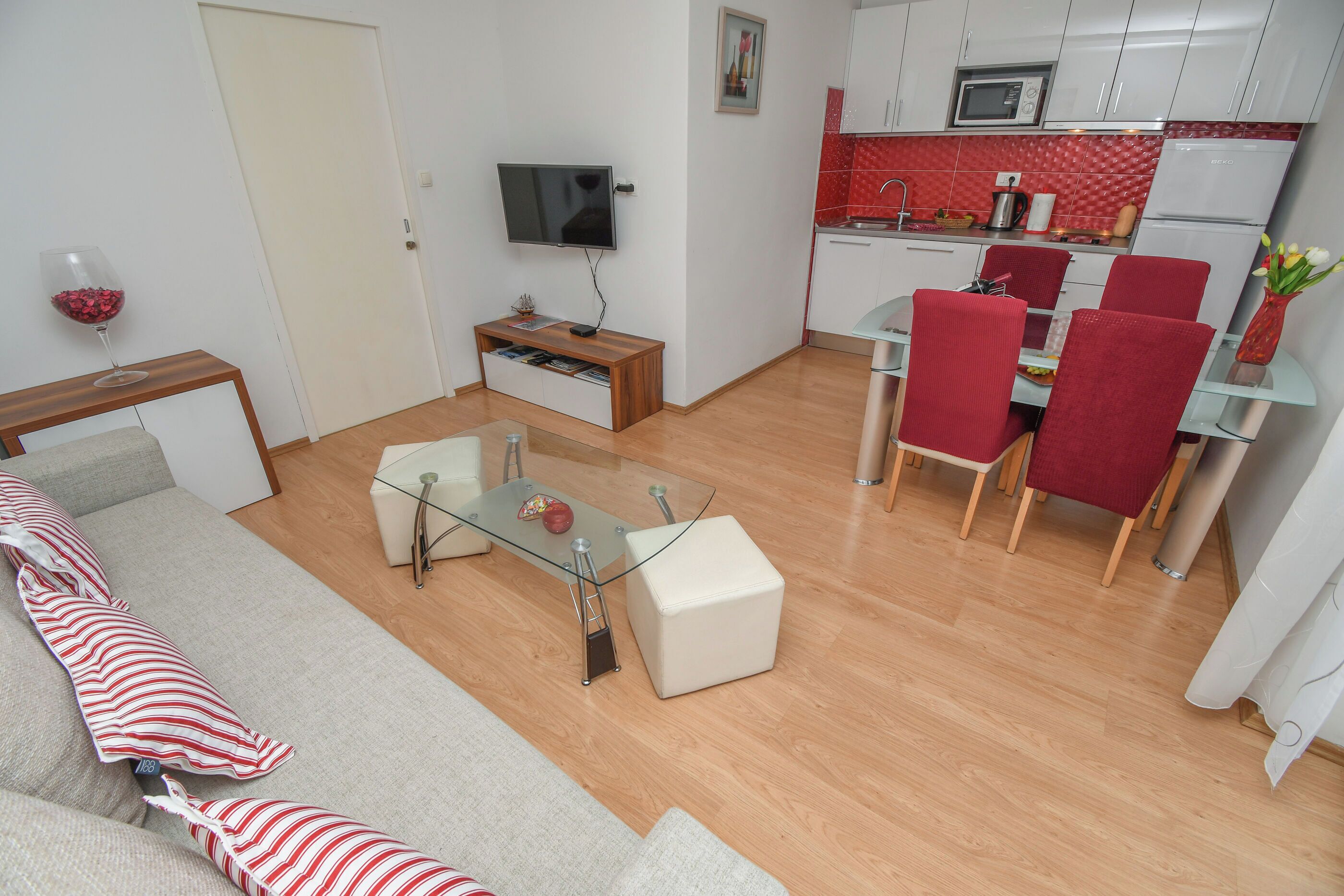 A modern living room with a glass coffee table, a sofa, and a TV. Adjacent is a compact kitchen with a microwave, sink, and red tiled backsplash.