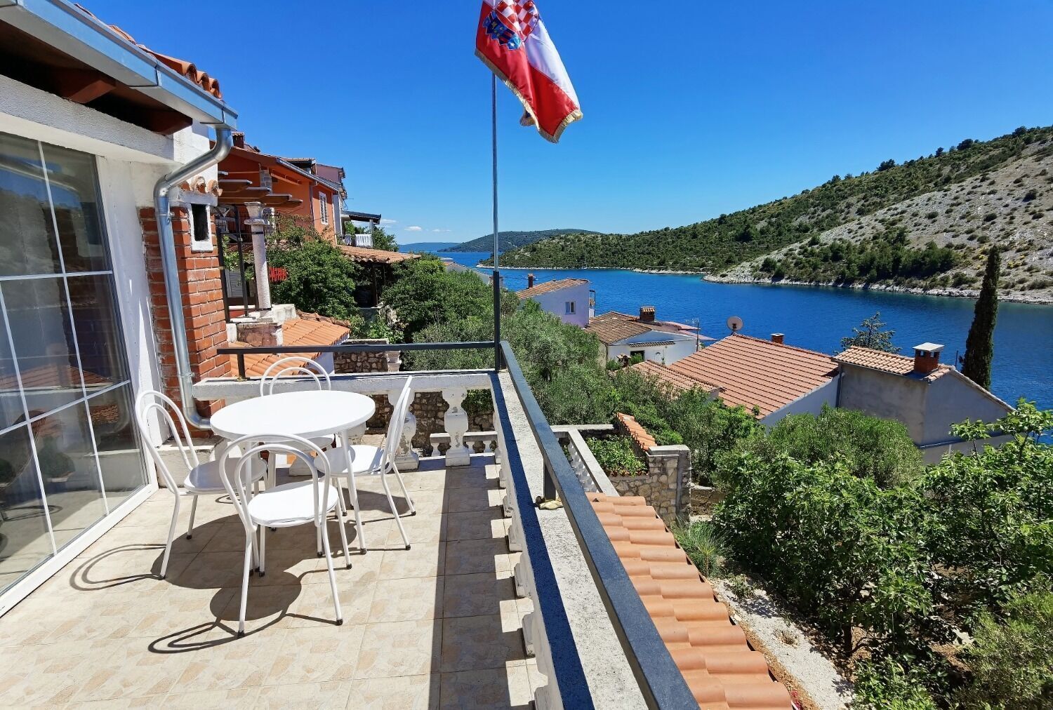 A balcony with a table and chairs overlooking a lake, a flag, and a view of the surrounding landscape.