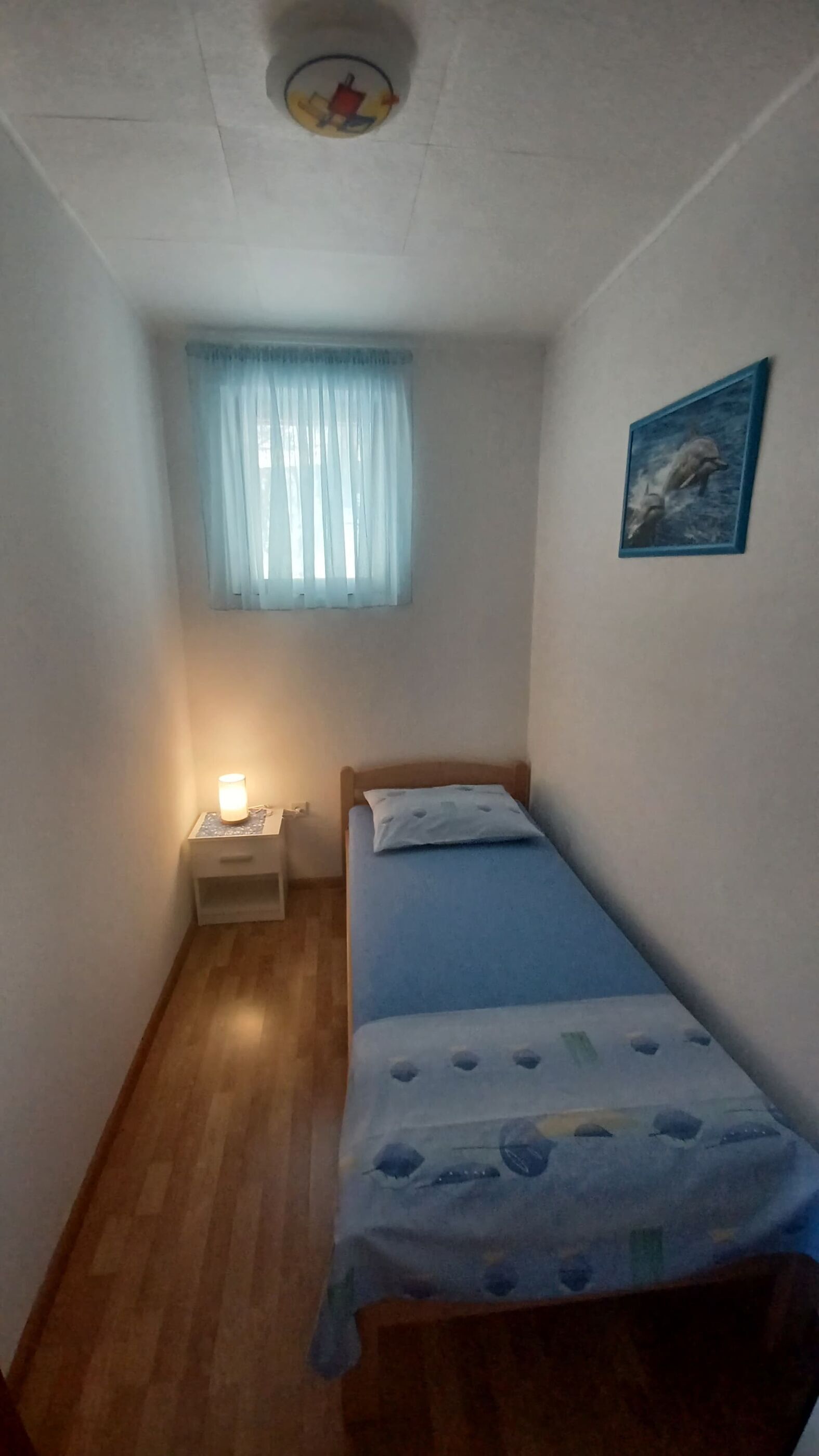 Een kleine, smalle kamer met een enkel bed, een nachtkastje, een raam met dunne gordijnen en een aan de muur hangend schilderij.