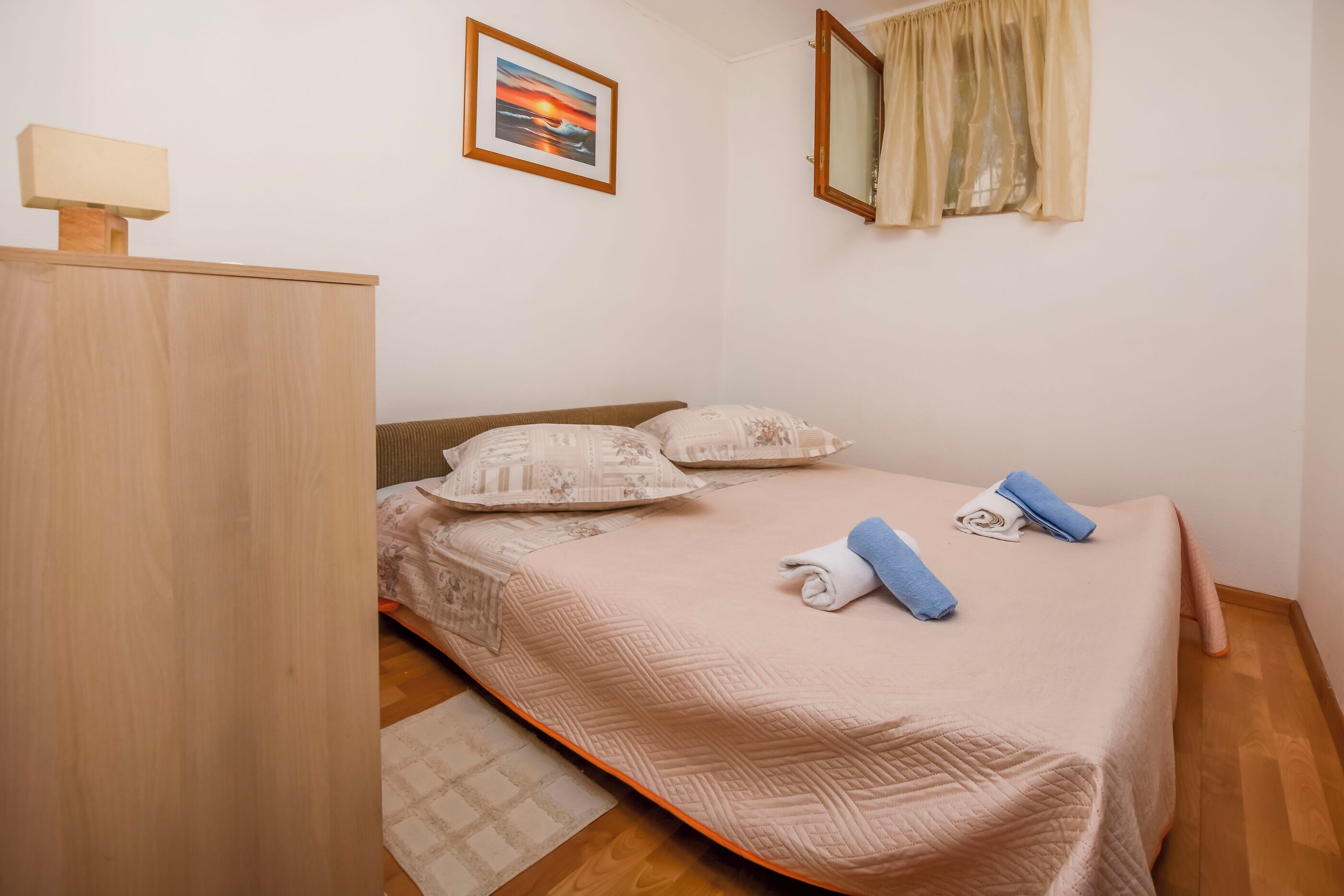 Une chambre à coucher avec un lit, des oreillers, une table de chevet, une lampe, une fenêtre avec des rideaux et un tableau au mur.