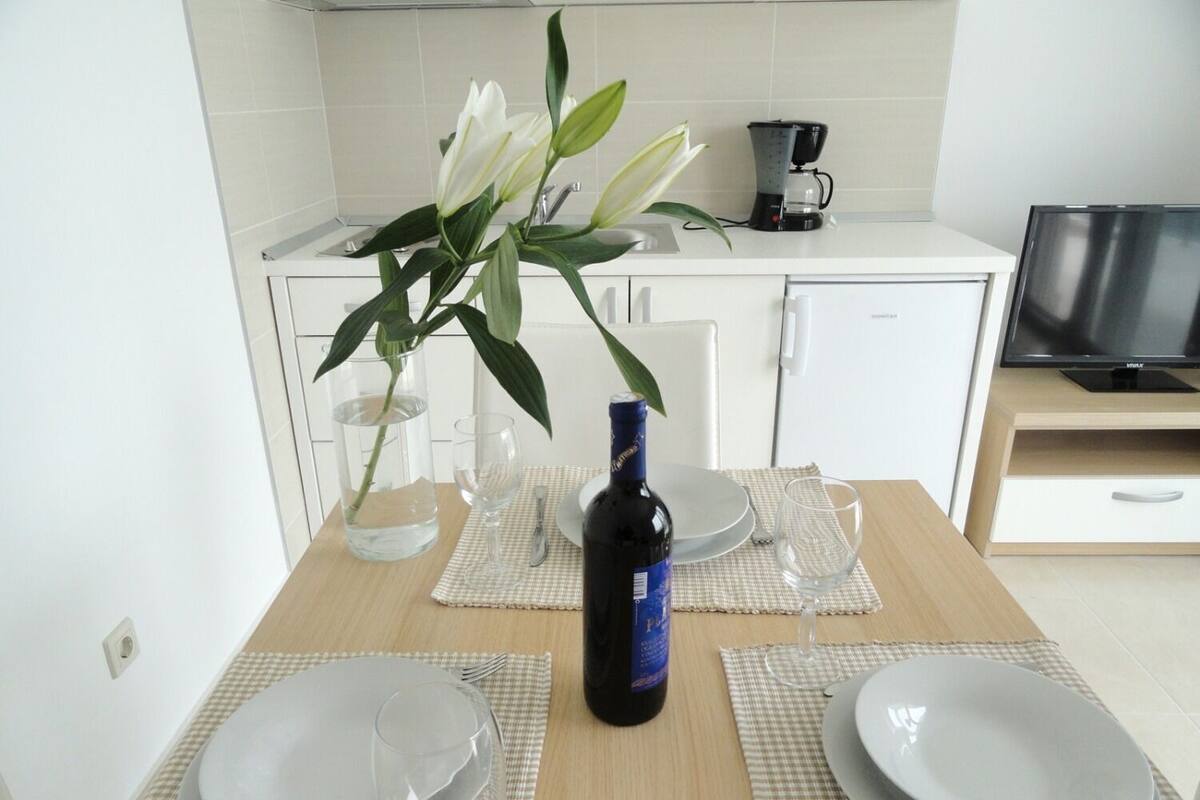 A modern kitchen with white cabinets, a coffee maker, a TV, and a dining table set with plates, glasses, and a bottle of wine.