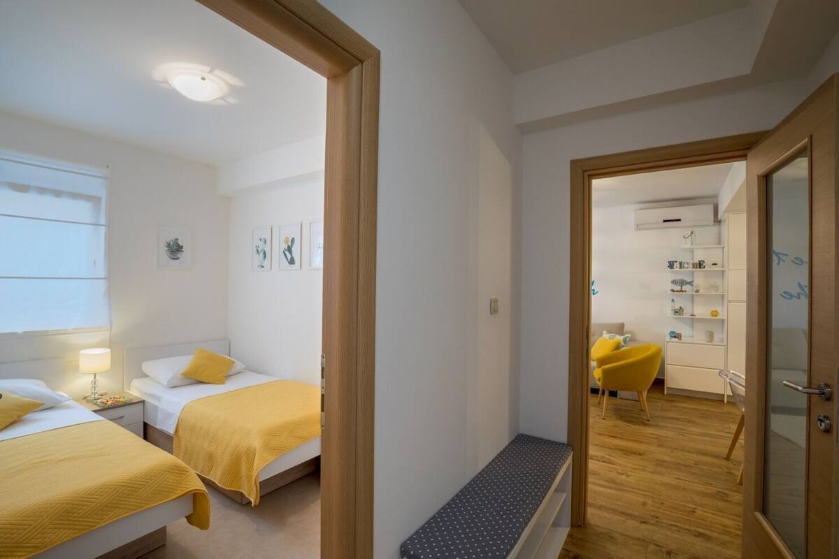 A modern hotel room with two beds, a bench, and a hallway leading to a living area with a chair and shelves.