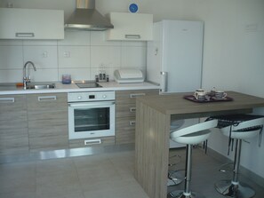 A modern kitchen with a built-in oven, a stainless steel range hood, a white refrigerator, and a bar area with stools. - Apartments Mirela (Omis)