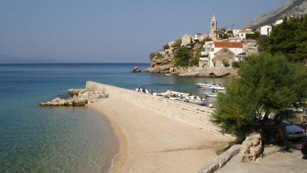 A coastal beach with clear waters, a stone staircase leading to a building with a tower, and several boats docked. - Apartments Mirela (Omis)