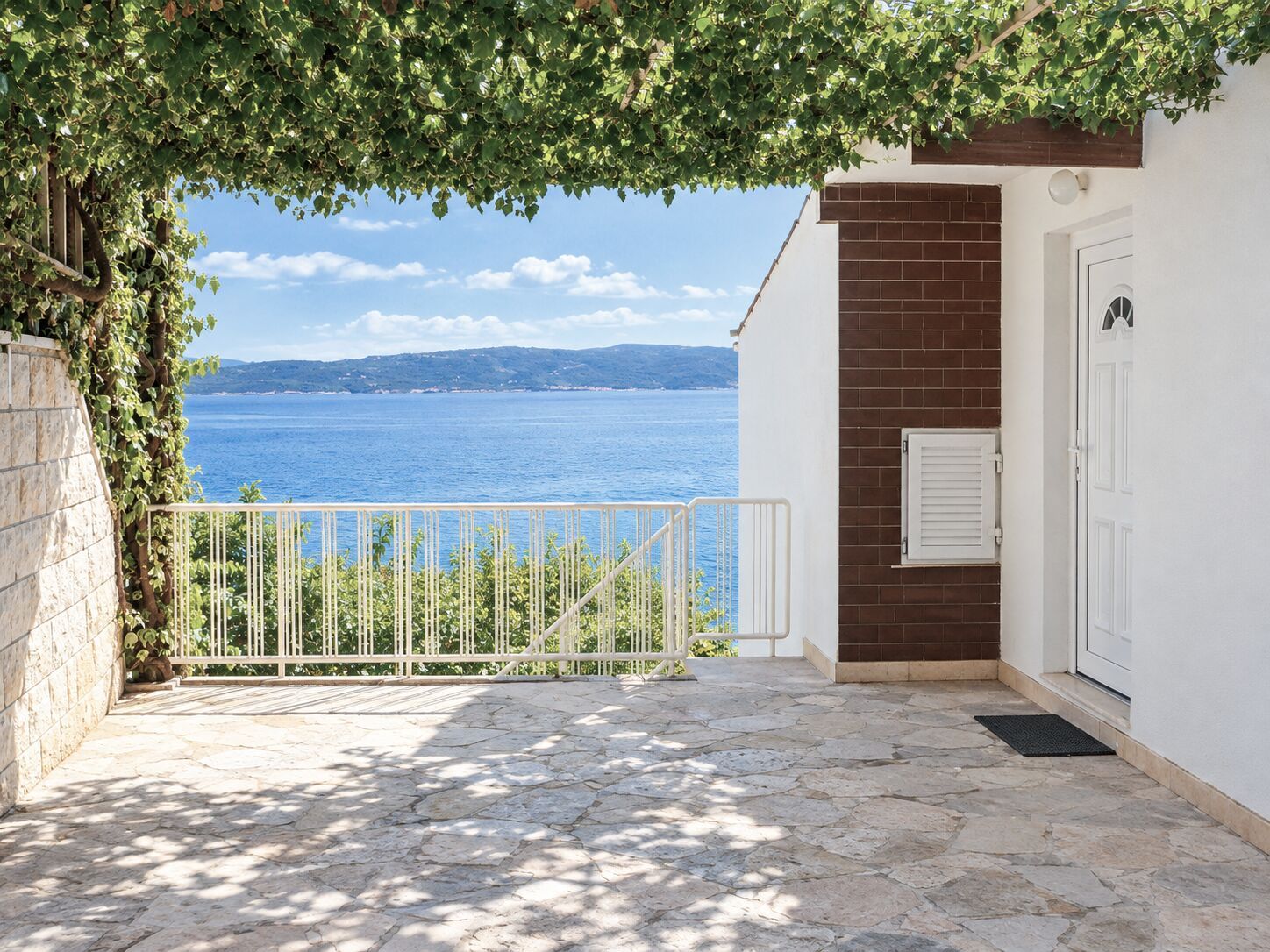 A stone-paved balcony with a white railing, overlooking a body of water and a distant coastline.