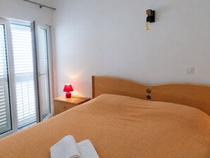 A bedroom with a bed, a nightstand with a lamp, a wall-mounted light, and a window with blinds. - Apartments Mirela (Omis)