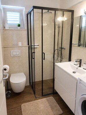 A modern bathroom with a glass-enclosed shower, a toilet, a sink, and a washing machine. - Apartments Mirela (Omis)