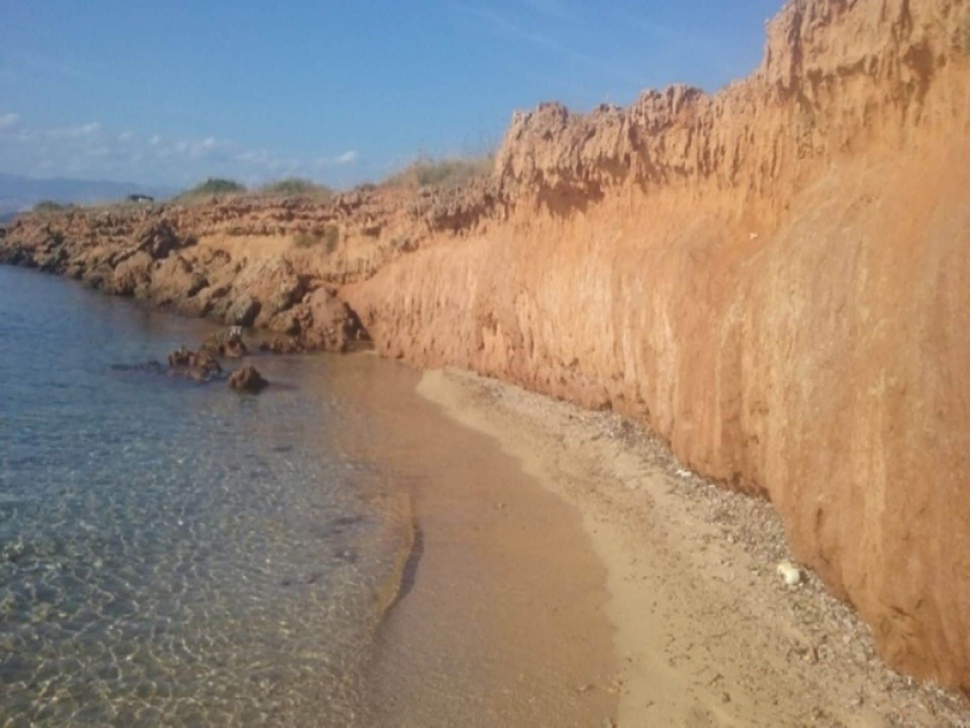 Uma praia costeira com águas rasas e cristalinas, falésias rochosas e vegetação rasteira.