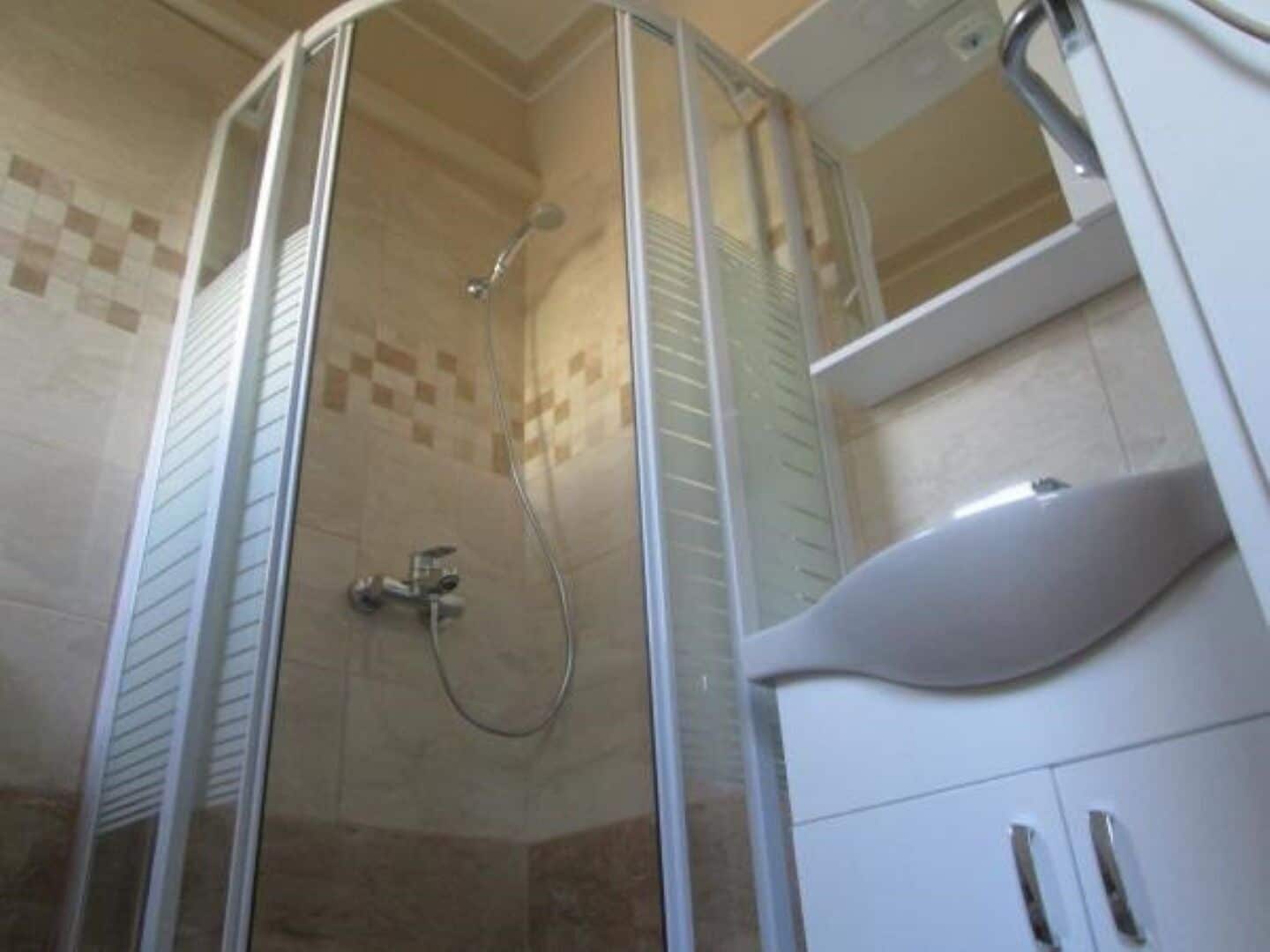 Area doccia vetrata con soffione a mano, lavandino bianco con lavabo curvo e parete rivestita di piastrelle a motivi a scacchi.