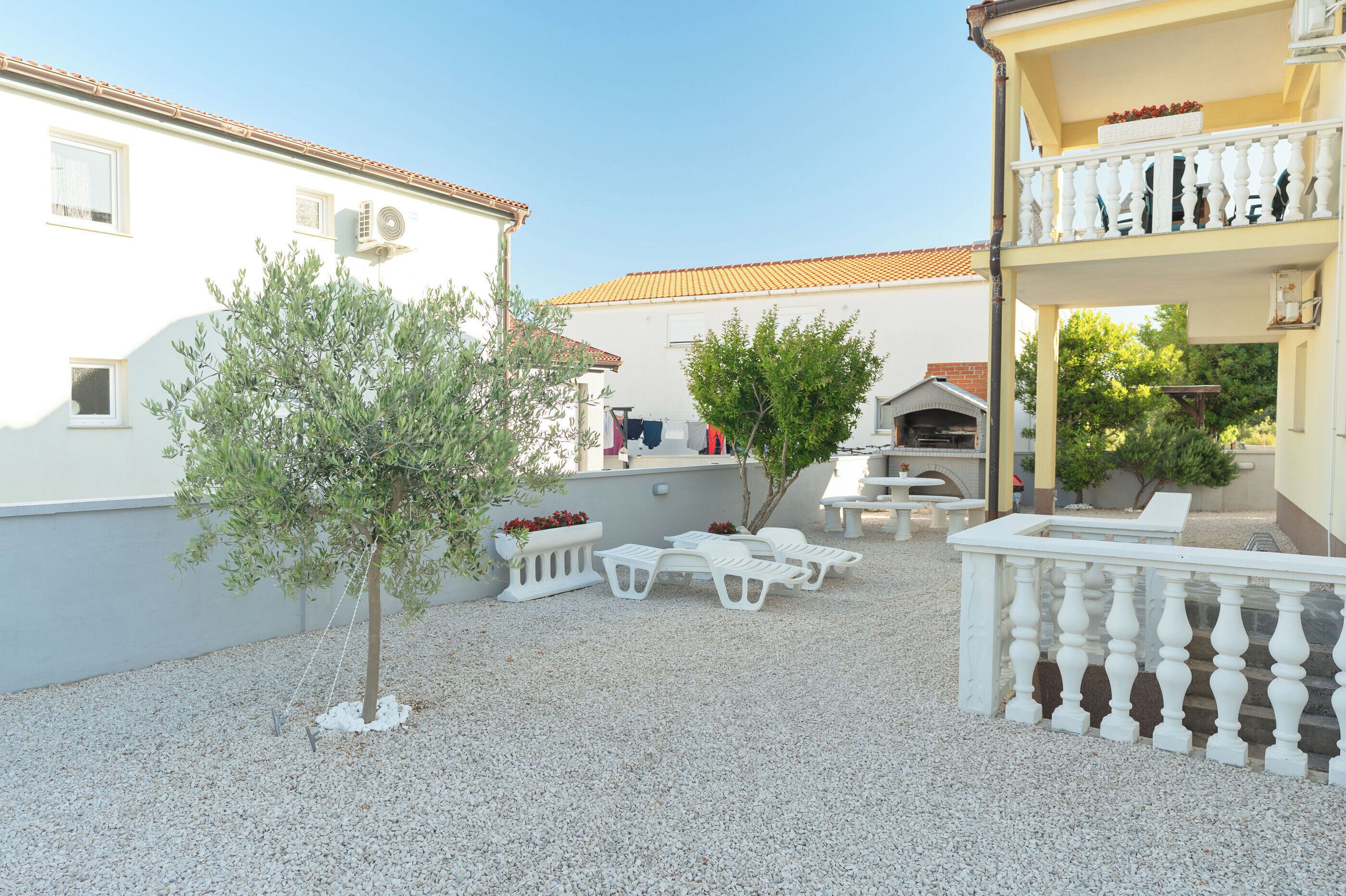 Un espace extérieur aménagé en patio, avec une clôture blanche, un petit arbre et des chaises en plastique blanches.