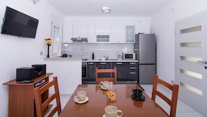 Una cocina compacta con armarios blancos, un microondas, un refrigerador y una mesa de comedor con un hervidor, tazas y un tarro de vidrio.
