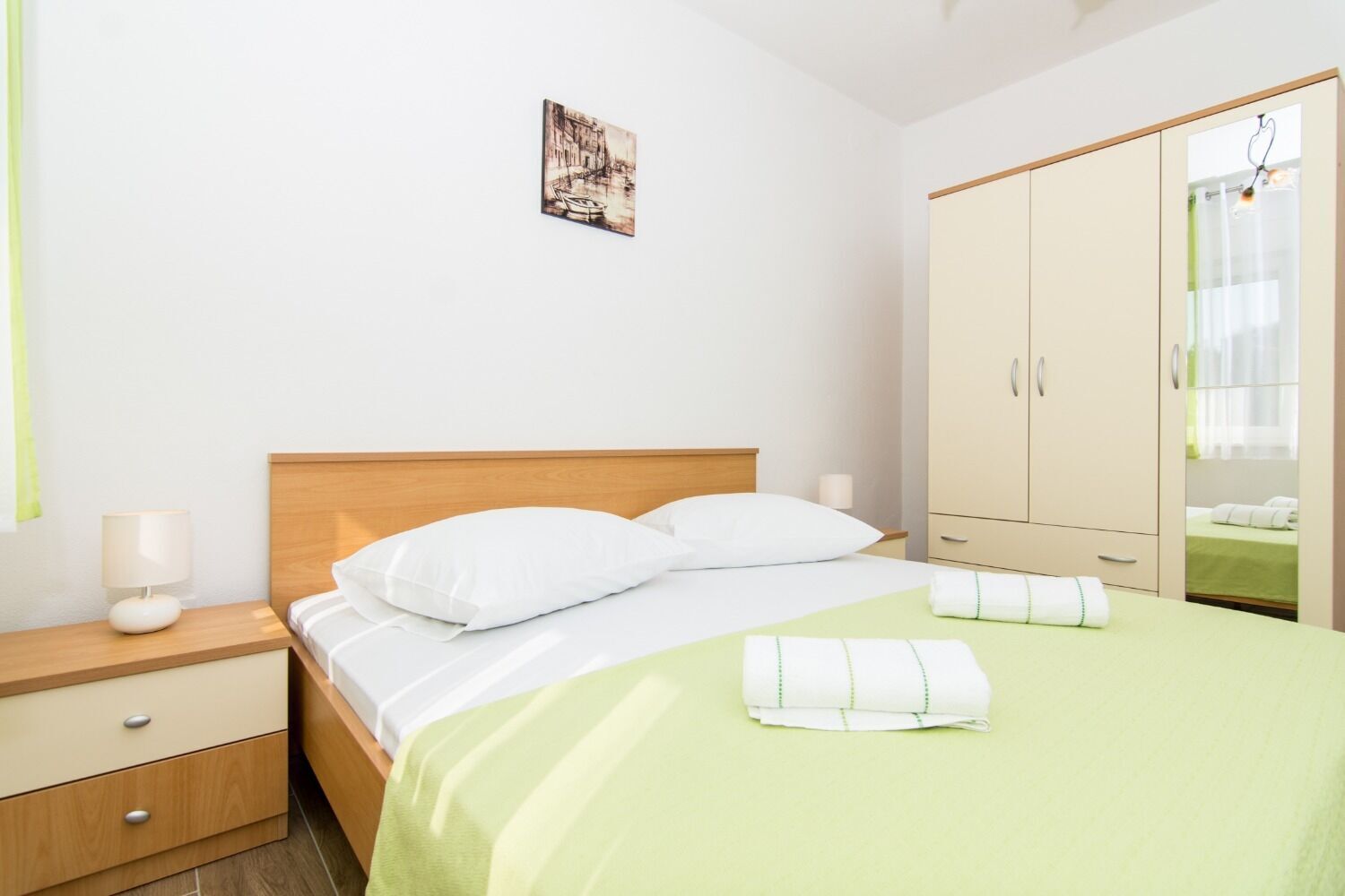 A neatly made bed with white linens, a wooden headboard, and a green bedspread. There is a nightstand with a lamp and a drawer. A large wardrobe with a mirror is to the right.