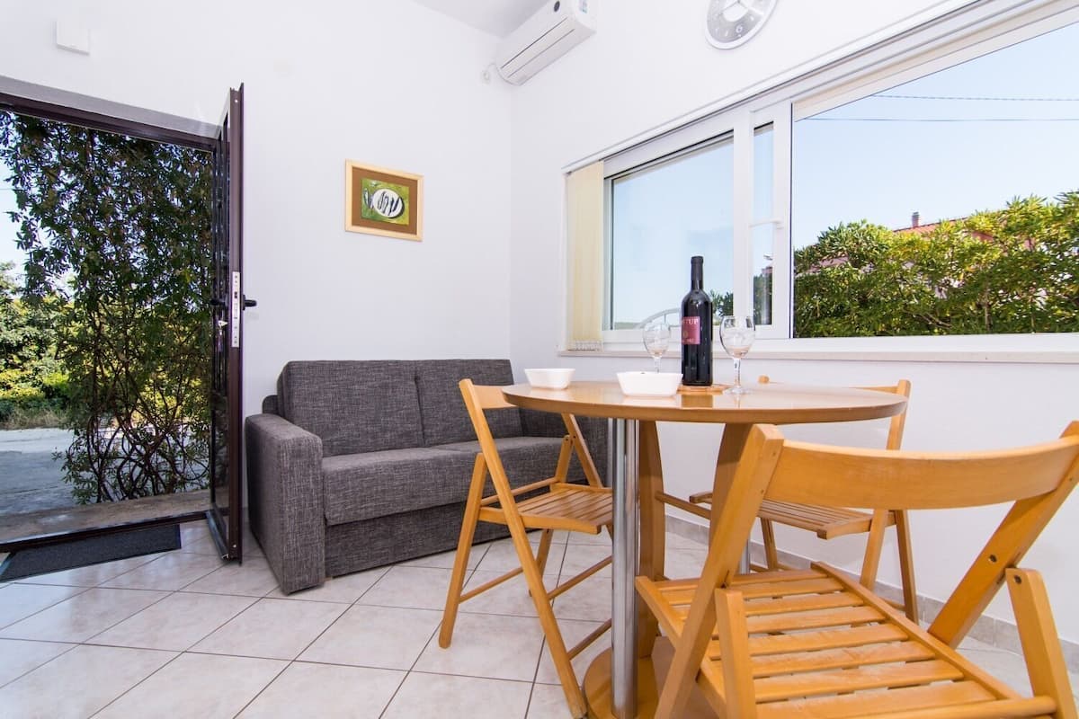 A small, modern room with a grey sofa, wooden chairs, a small table, and a bottle of wine.