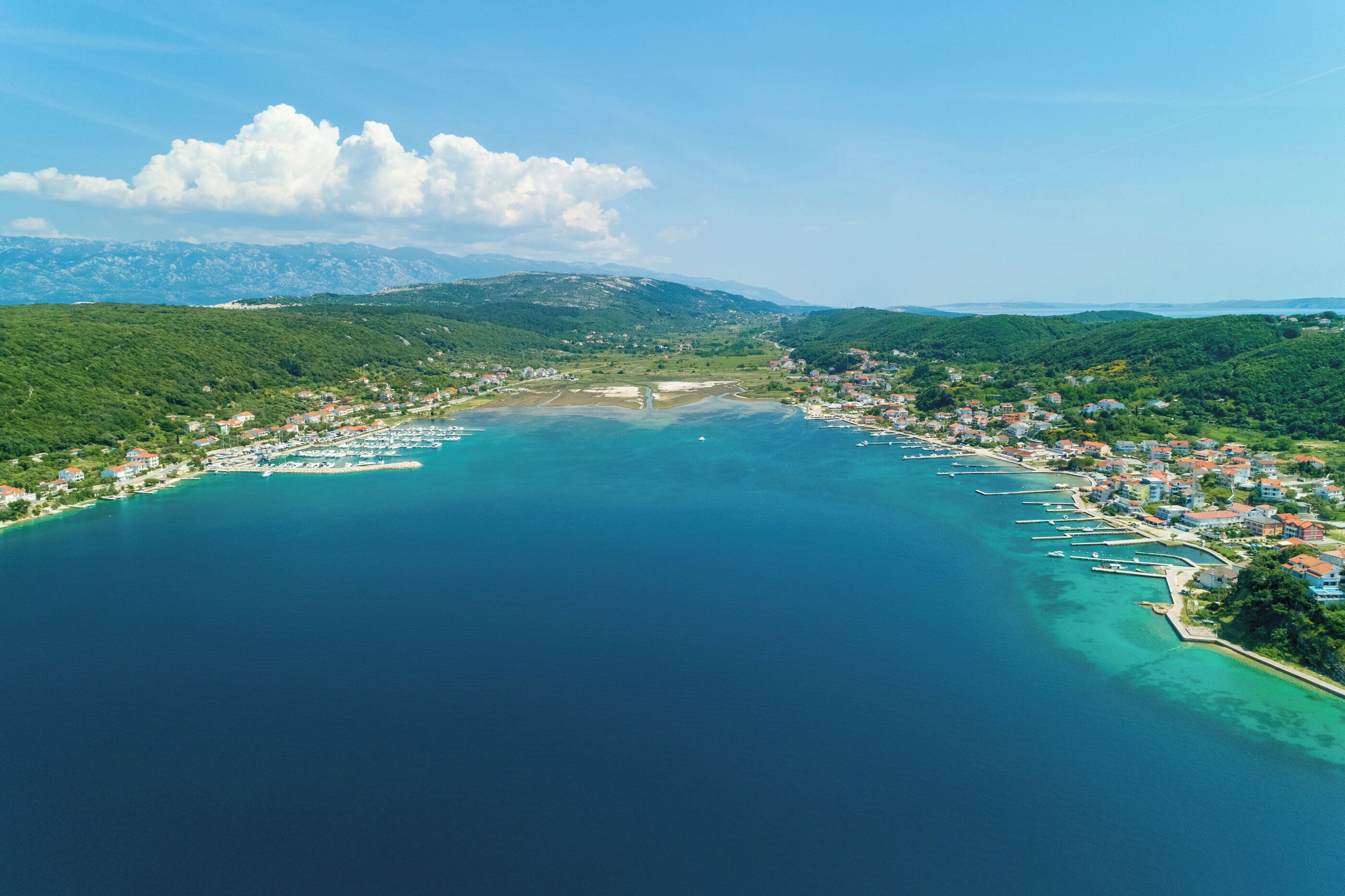 An aerial view of a coastal town with a marina, surrounded by green hills and mountains in the background.