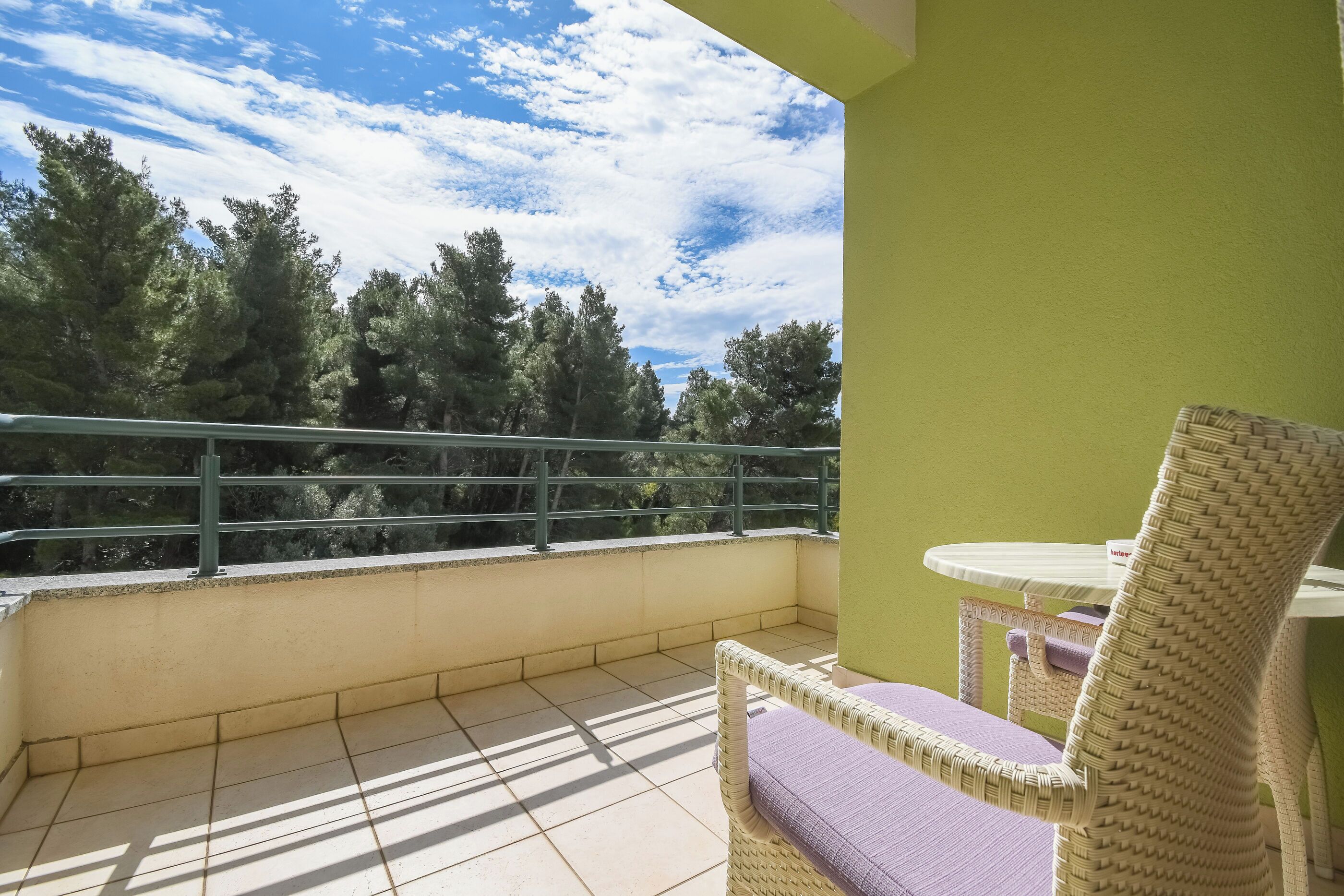 Ein Balkon mit Korbstuhl und kleinem Tisch, mit Blick auf eine bewaldete Landschaft.