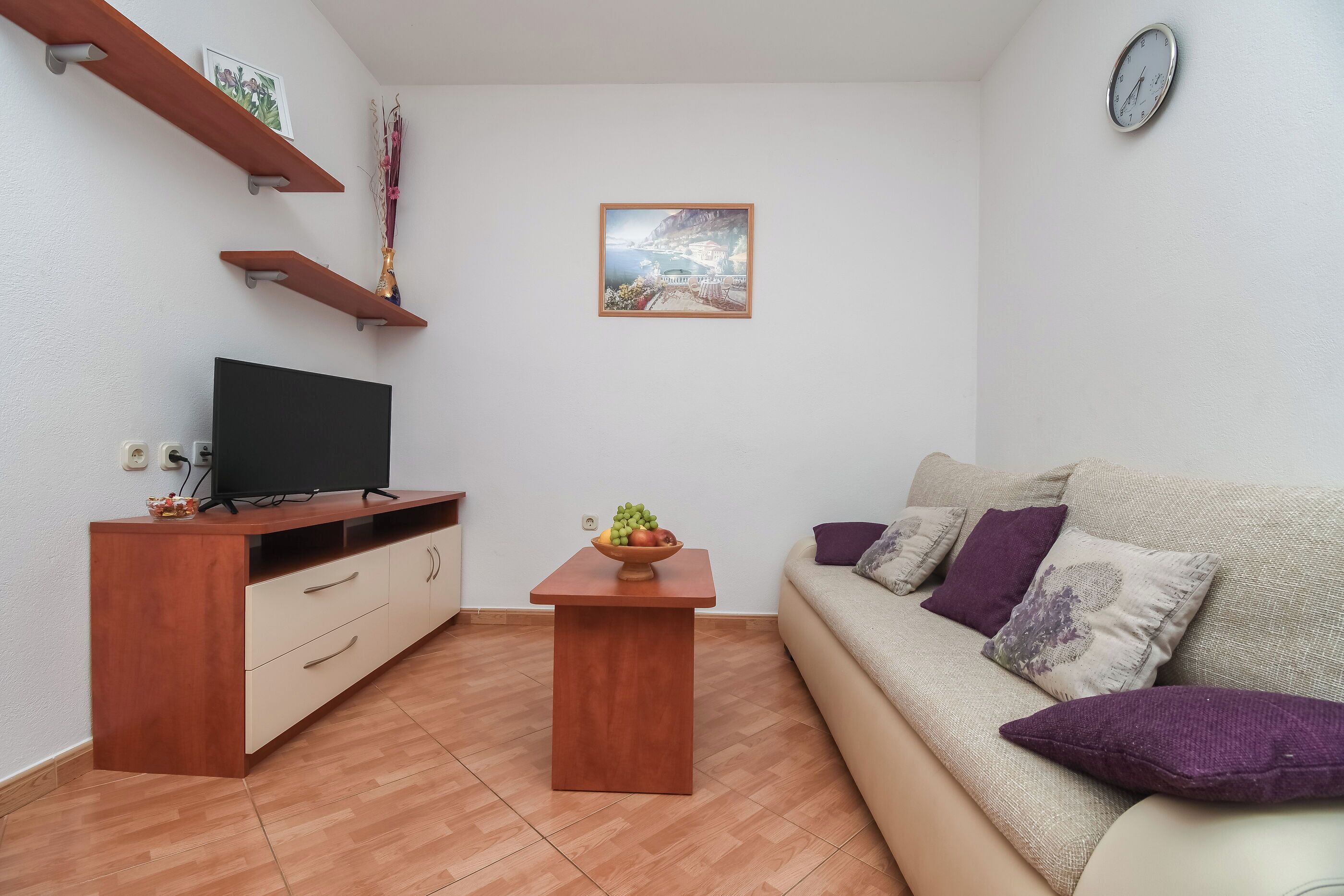 A living room with a TV, wooden shelves, a small table with fruit, and a sofa with cushions.